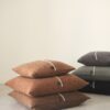 Seis cojines de diseño en lana merino apilados en dos montones. En el primer montón cojines en tonos anaranjadas en el segundo montón colores gris, verde y negro