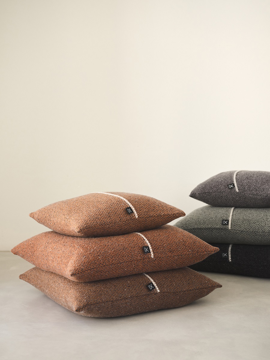 Seis cojines de diseño en lana merino apilados en dos montones. En el primer montón cojines en tonos anranjadas en el segundo monton colores gris, verde y negro