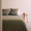 Cojín y manta de lana merino con trazabilidad diseño en cuadrados verdosos, amarillos y azules con perfiles en amarillo expuestos sobre cama