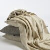Manta de lana merino tejida en telar manual con trazabilidad en colores neutros con perfiles internos en gris apoyada sobre cojines con misma composición y diseño