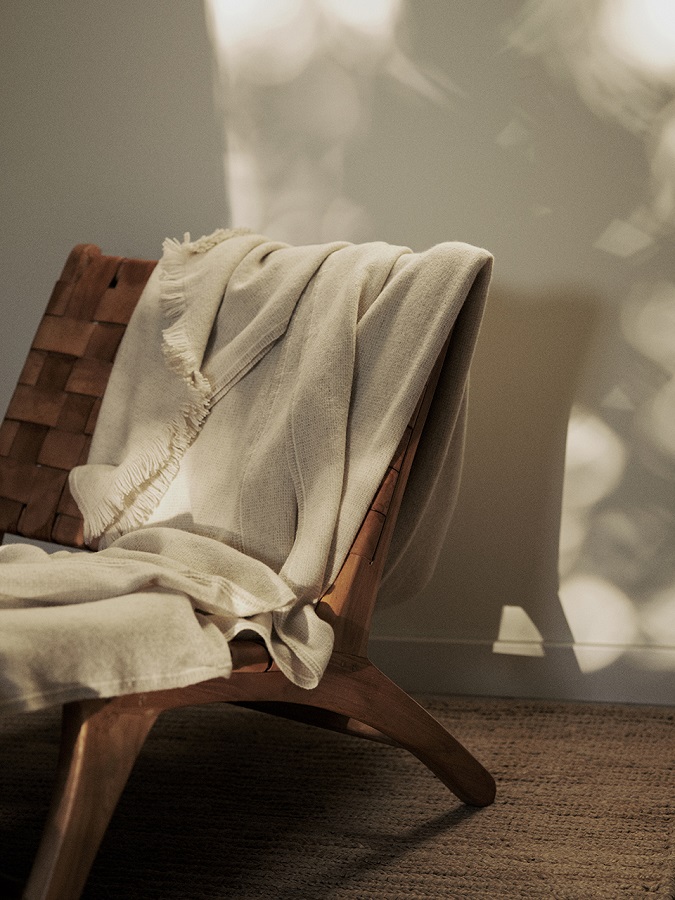 Manta de diseño de Teixidors Nebula en lana cashmere y merina en tono crudo apoyada sobre butaca en tonos piel oscura